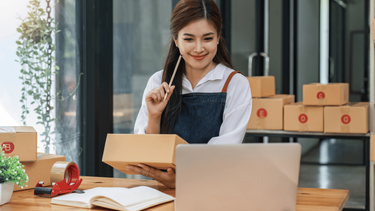 A woman checking her laptop while preparing shipment boxes for selling on Amazon with GS1 barcodes.