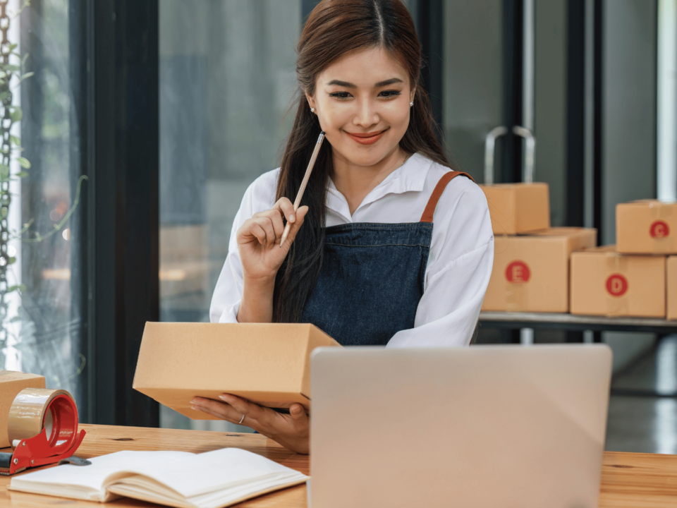 A woman checking her laptop while preparing shipment boxes for selling on Amazon with GS1 barcodes.