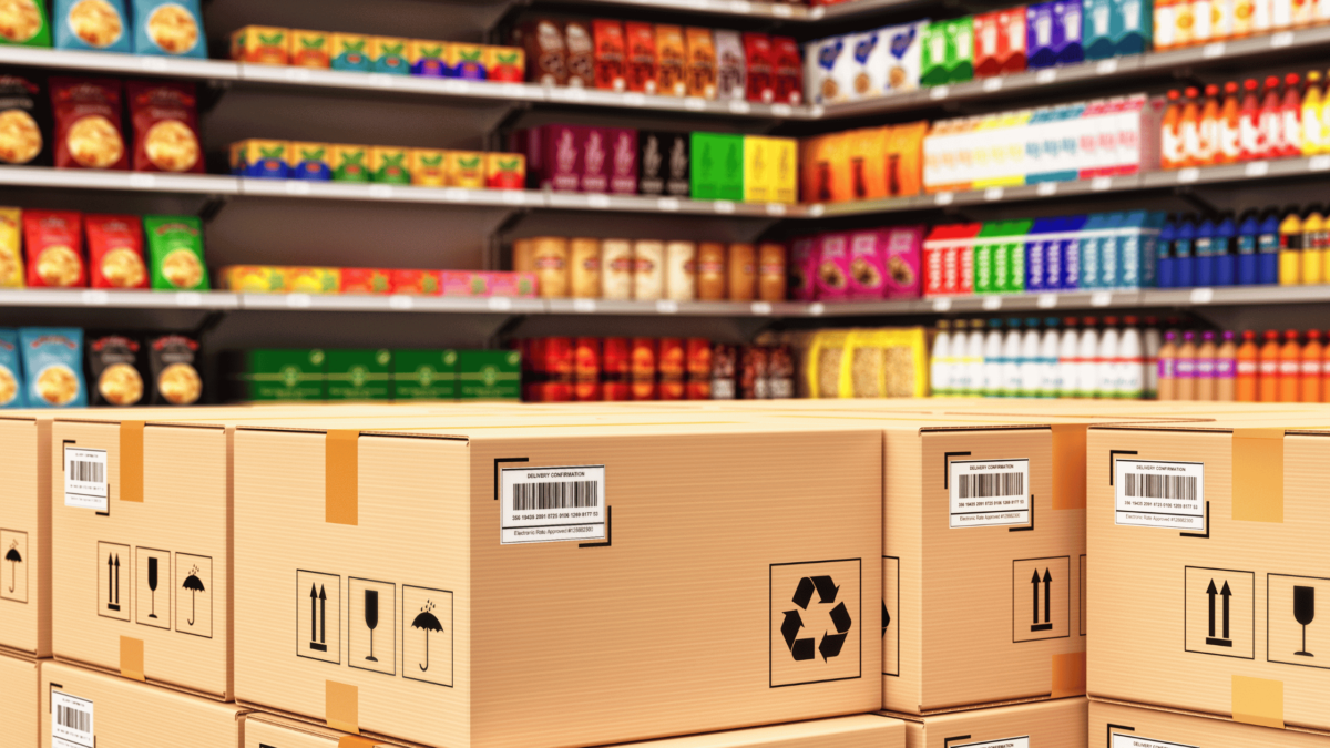 Stacked cardboard boxes with GS1 barcodes in a retail store aisle filled with colorful packaged products.