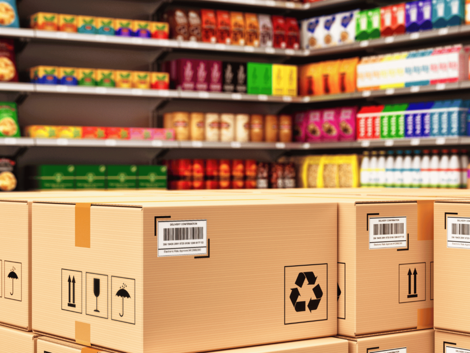 Stacked cardboard boxes with GS1 barcodes in a retail store aisle filled with colorful packaged products.