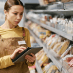 A person wearing an apron and a yellow shirt holds a tablet and checks inventory on shelves stocked with packaged goods in determining "how to format GS1 128 labels for food distribution."