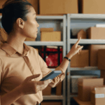 A person holds a phone and points to shelves stocked with cardboard boxes moving through the supply chain for "What is a GS1-128 Barcode."