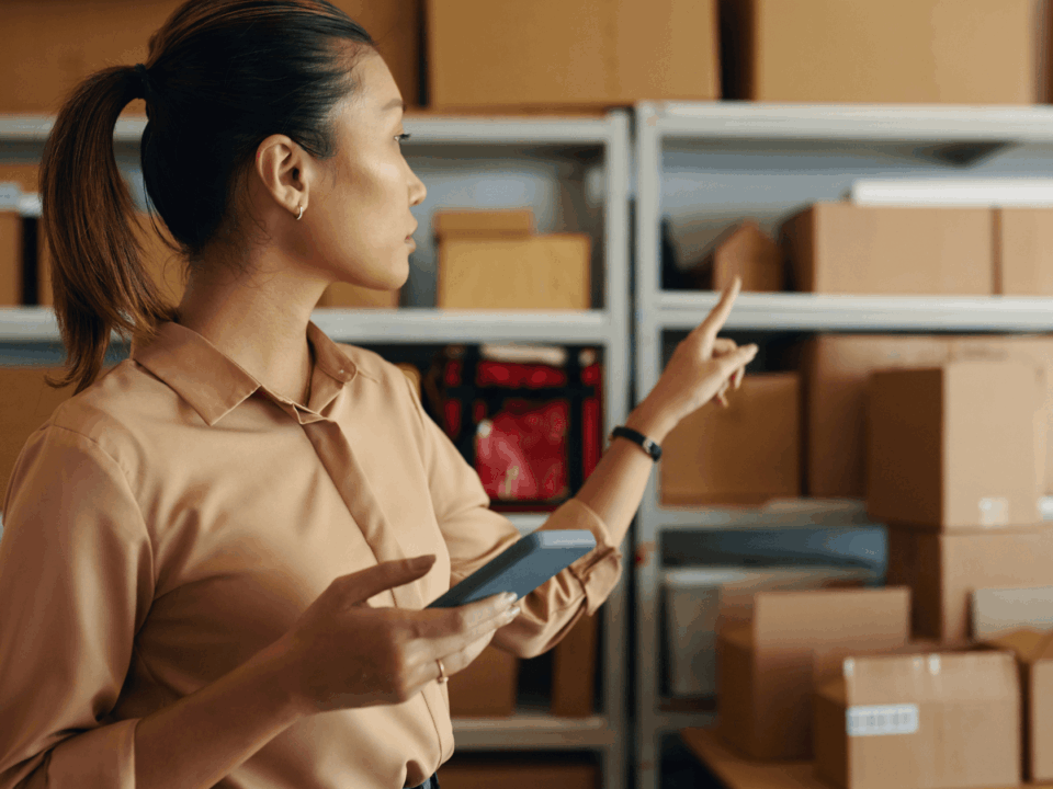 A person holds a phone and points to shelves stocked with cardboard boxes moving through the supply chain for "What is a GS1-128 Barcode."