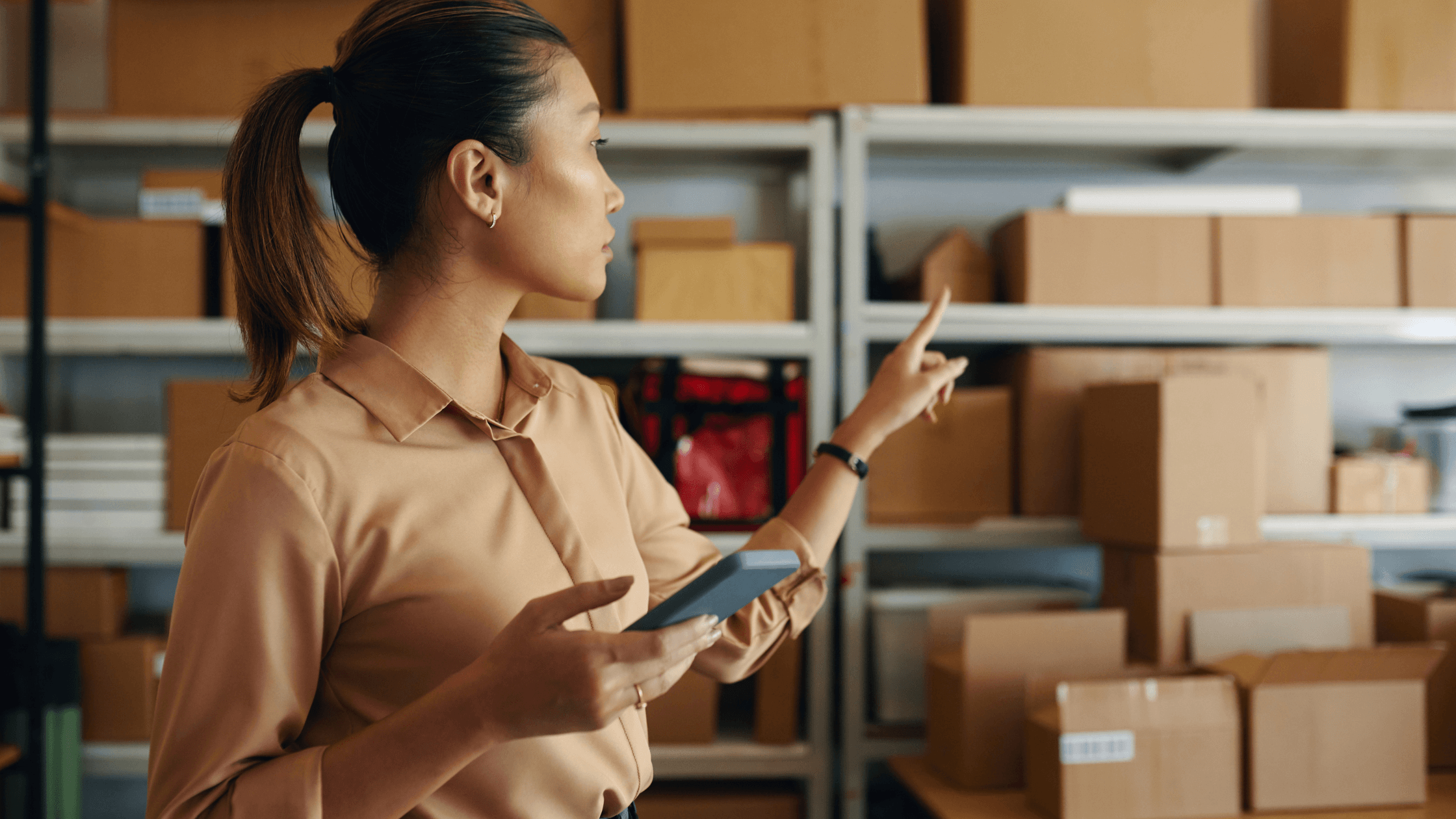 A person holds a phone and points to shelves stocked with cardboard boxes moving through the supply chain for "What is a GS1-128 Barcode."